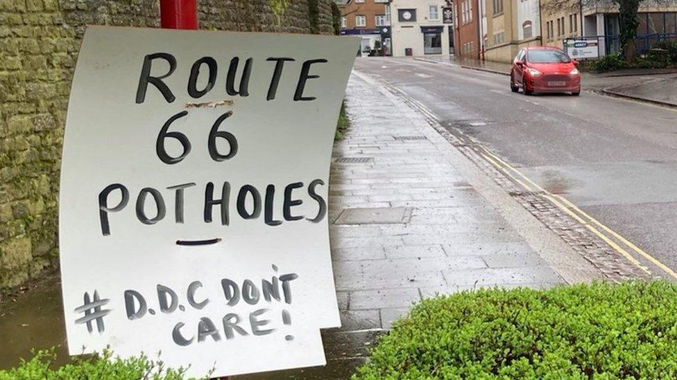 White sign with black lettering saying "Route 66 Potholes" on urban road