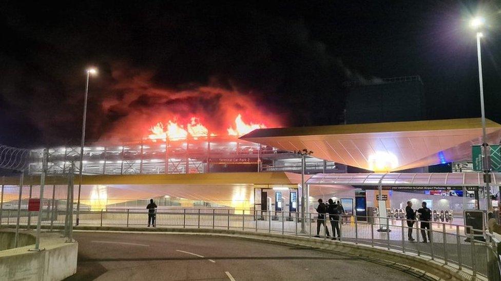 Flames visible on the roof of an airport car park. Some passengers are visible standing on the airport roadway.