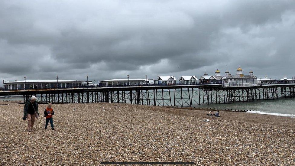 Empty pebbled beach with a cloudy, ominous, sky. Some people walking along the sea side. An old wooden dock in the background.