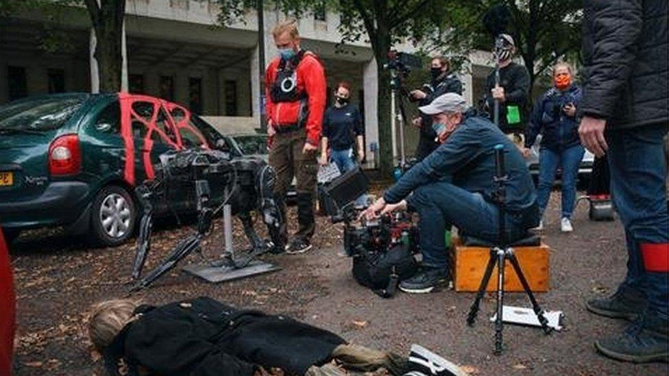 An actor lies on the floor in a chaotic street scene, with the camera operator and crew wearing masks