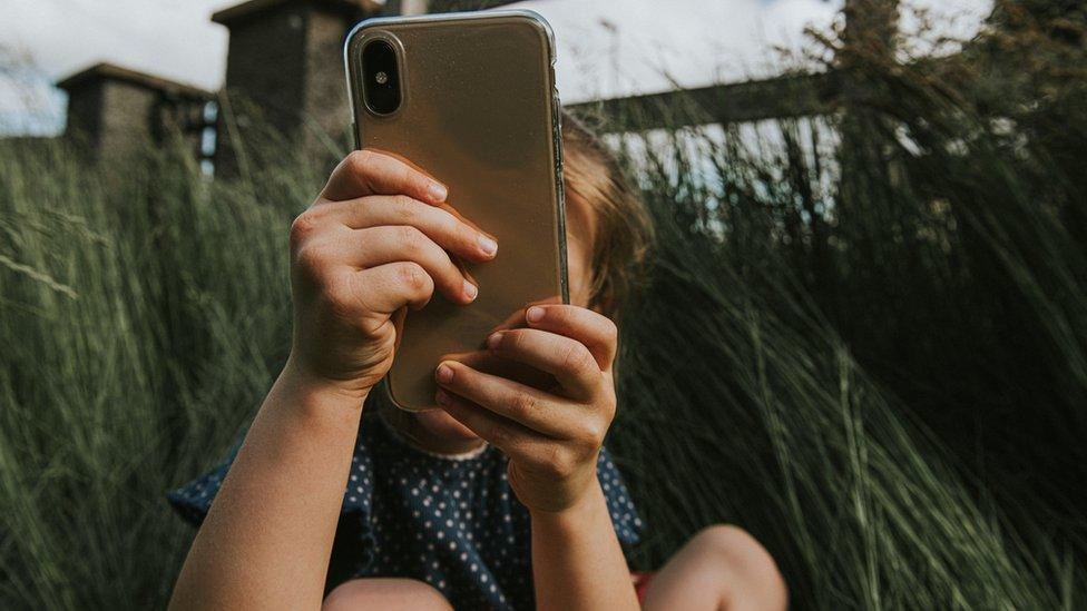 Young girl holds smart phone in front of her face taking a picture outside.