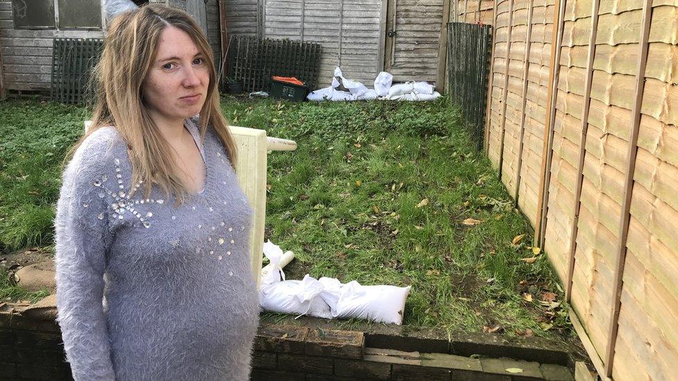 A heavily pregnant woman in her muddy garden with sandbags at the gate