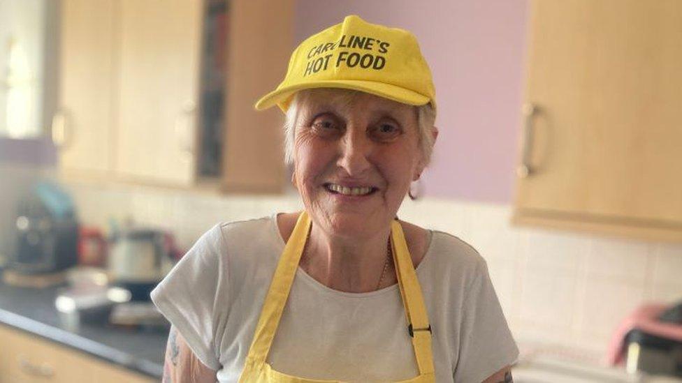 Caroline Dove in a yellow apron and hat that reads Caroline's Hot Food.