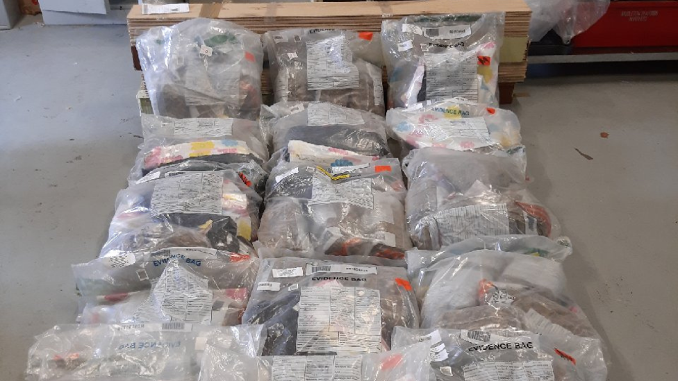 An image of several large clear evidence bags full of cannabis. The bags are lying on a concrete floor and are laying against some of the seized floorboards.
