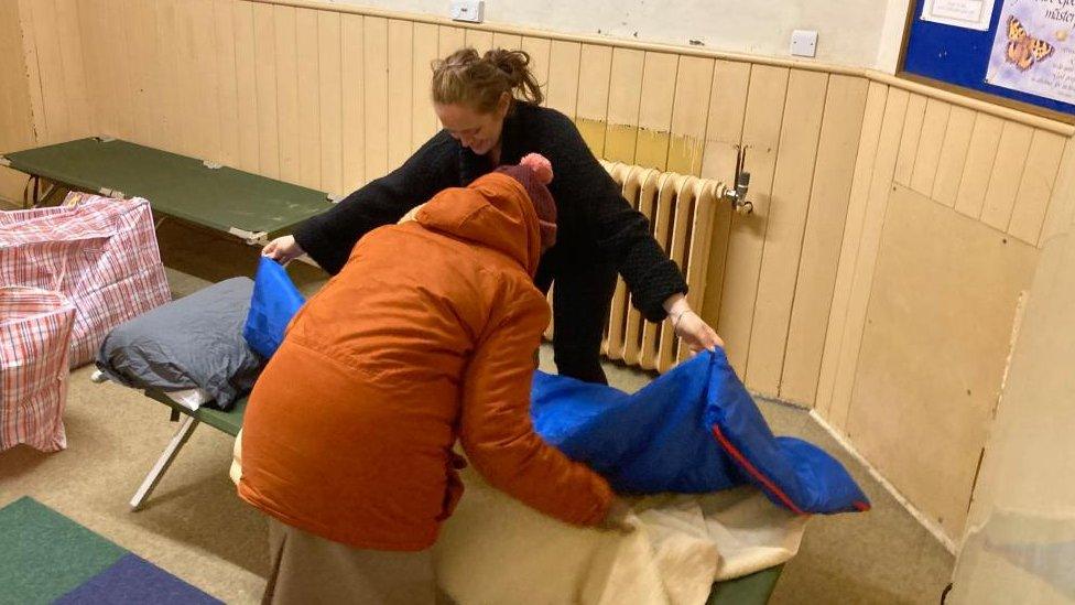 Woman in a raincoat arranges a blue sheet on a campbed with help from a Hope Centre staff member