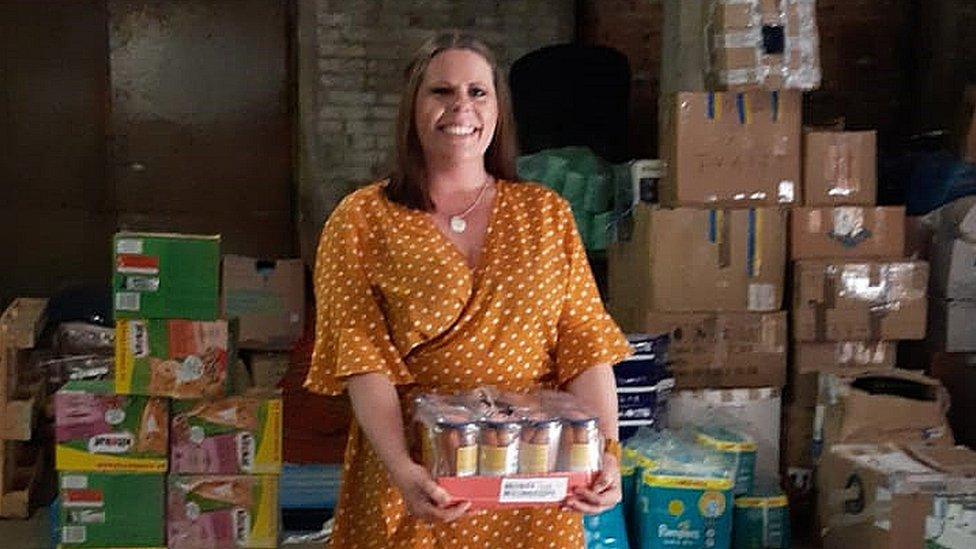 Lucie Shanks holding a pack of canned food in front of other supplies donated for refugees