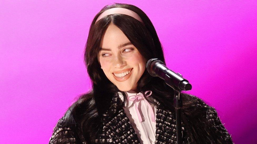 Billie Eilish performs onstage at the 96th Annual Oscars held at Dolby Theatre on March 10, 2024 in Los Angeles. Billie is a 22-year-old white woman with long black hair and green eyes. She wears a baby pink headband, matching a pink ribbon bow on her white shirt. She wears a black and white overcoat and smiles, looking to the right. She stands in front of a microphone and the stage behind her is lit a vibrate pink