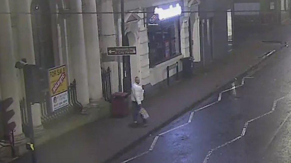 A man wearing a light-coloured jacket holding a shopping bag walking on the pavement on Bedminster Parade