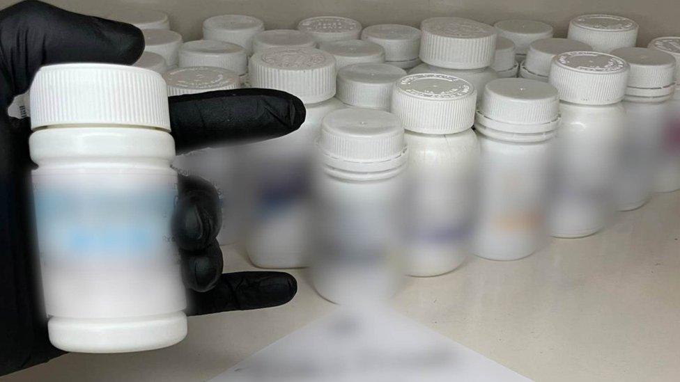 Black glove holding bottle of pills with more bottles in the background