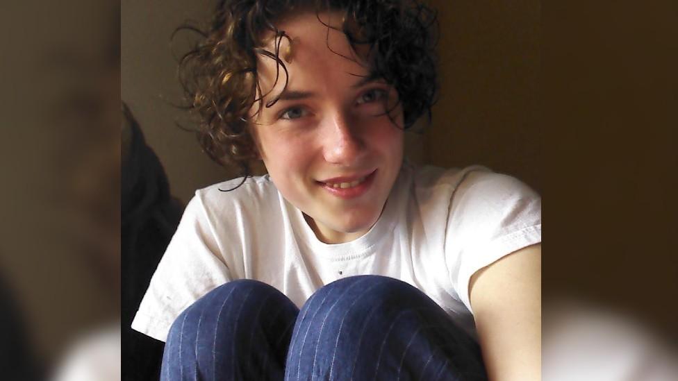 Evangeline Wilson, with curly hair, wearing a white t-shirt and blue trousers.