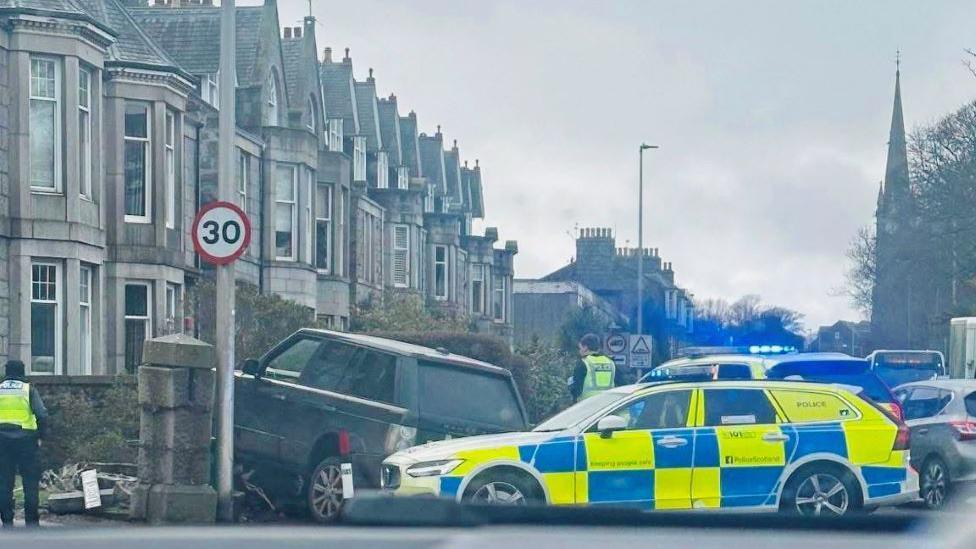 Accident scene with a Range Rover crashed through a garden wall, with blue and yellow-checked police cars, and traffic, and rows of houses, and a church spire in the background.