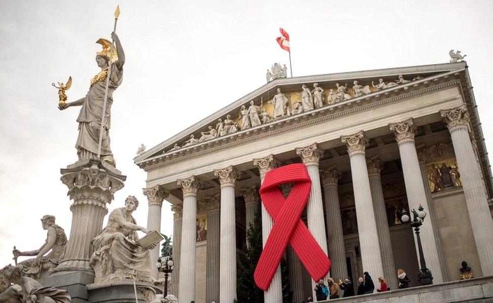 People stand in front of a symbolic "Red Ribbon" attached to the facade of the Austrian parliament building