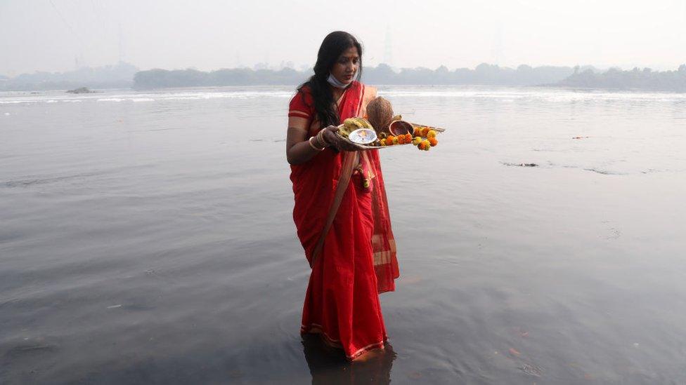 A woman dressed in traditional clothing makes an offering while standing in the river