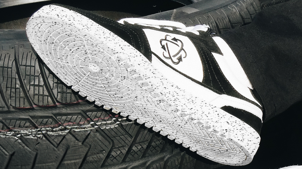 A close-up of one of the trainers in front of a tyre. The shoe is black and white, with a speckled sole.