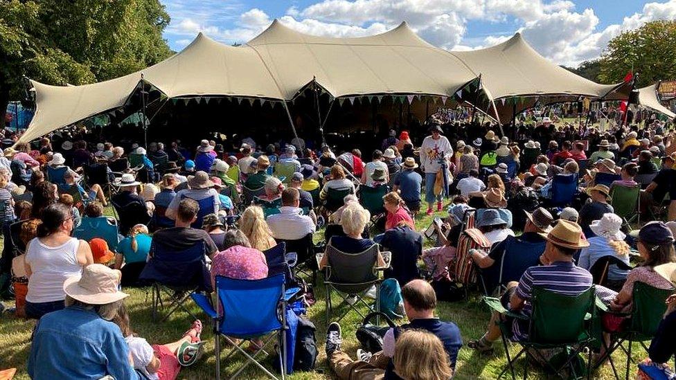 A crowd of people sitting on the grass outside a marquee