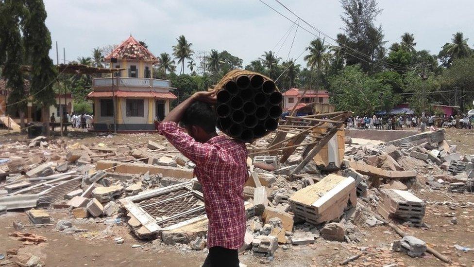 A man walks carrying empty shells of fireworks past a collapsed building after a massive fire broke out during a fireworks display at the Puttingal temple complex in Paravoor village, north of Thiruvananthapuram, southern Kerala state, India,