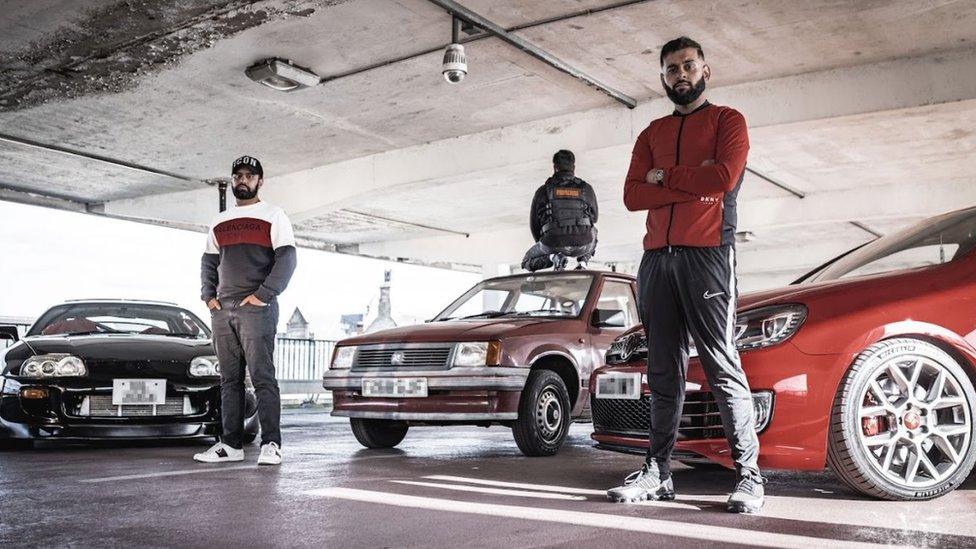 A publicity shot for Peaceophobia. Two bearded men are standing in a car park, one with his hands in his pockets, the other his arms folded. Behind them are three parked cars, facing them. A third man, facing away from the camera, is crouched on top of the car in the middle.