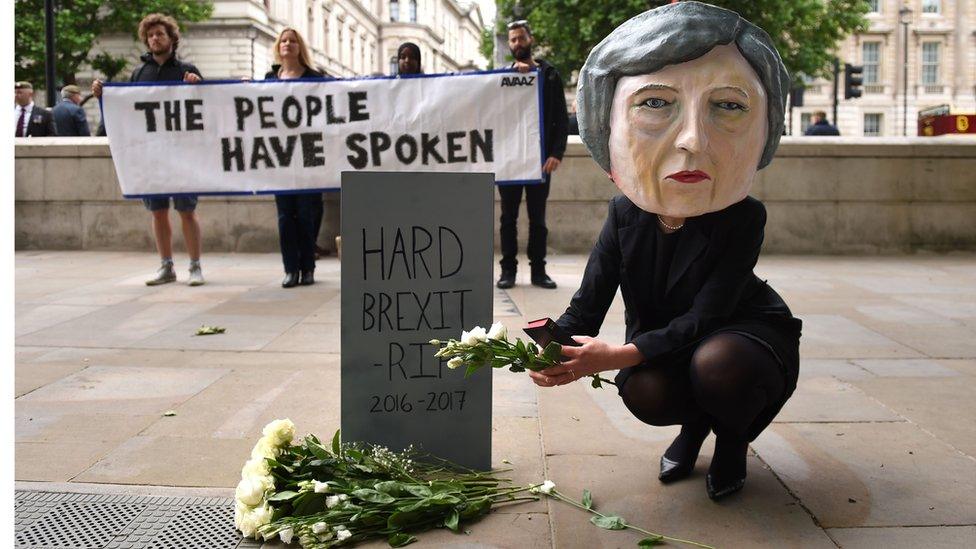 A life-size giant-headed puppet of Theresa May leaving flowers at a tombstone bearing the words Hard Brexit RIP