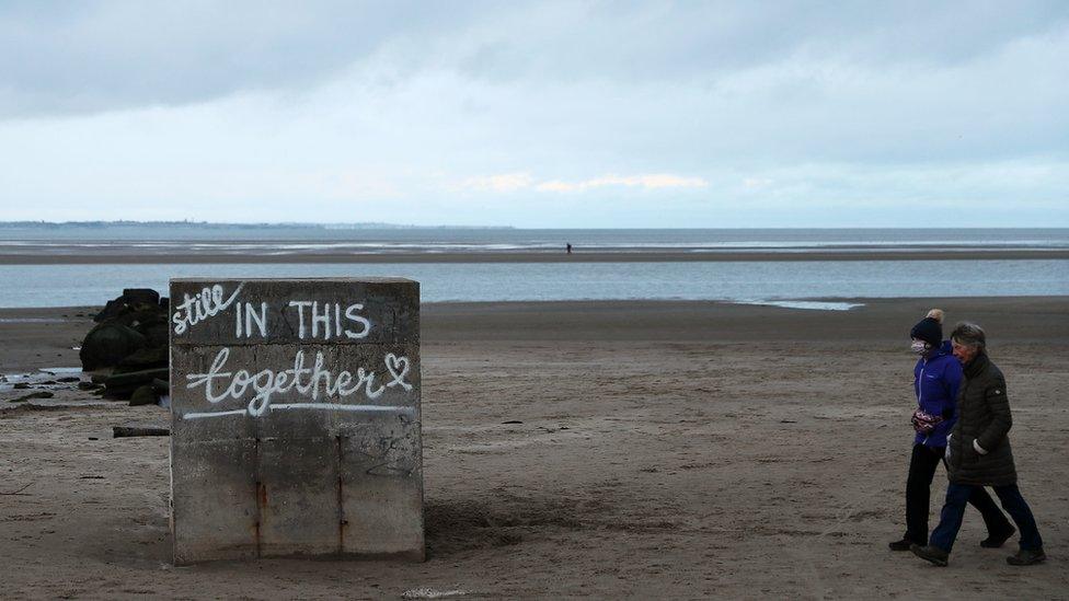 A man and a woman walk along a beach in Dublin where the words "Still in this together" have been painted on a concrete slab