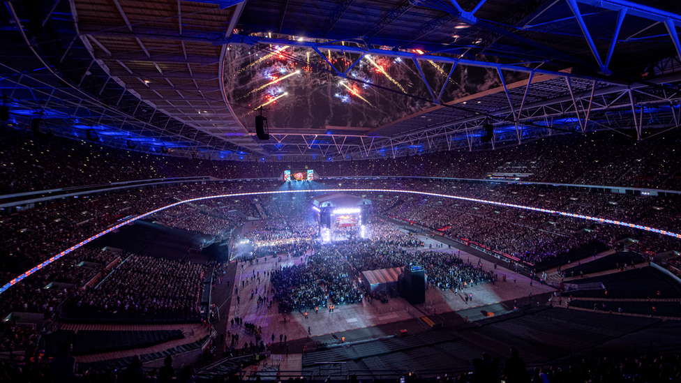 Crowds gather for the All Elite Wrestling event in Wembley Stadium. The stadium is seen from a high angle looking across the majority of the venue. Fans are seated around the stadium which has a stage and a crowd in the middle. It is night time and red and yellow fireworks are being lit from the roof. The stadium is lit by purple-coloured lights.