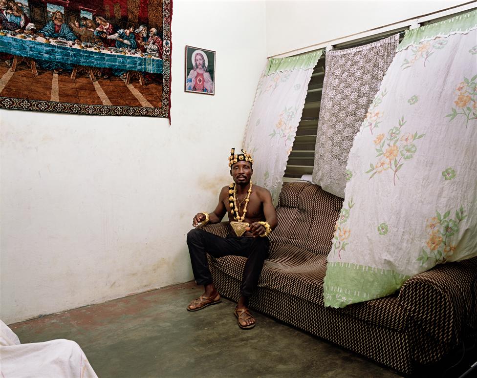 A photo by Deana Lawson of a shirtless man sitting on a sofa, with religious images beside him on the wall