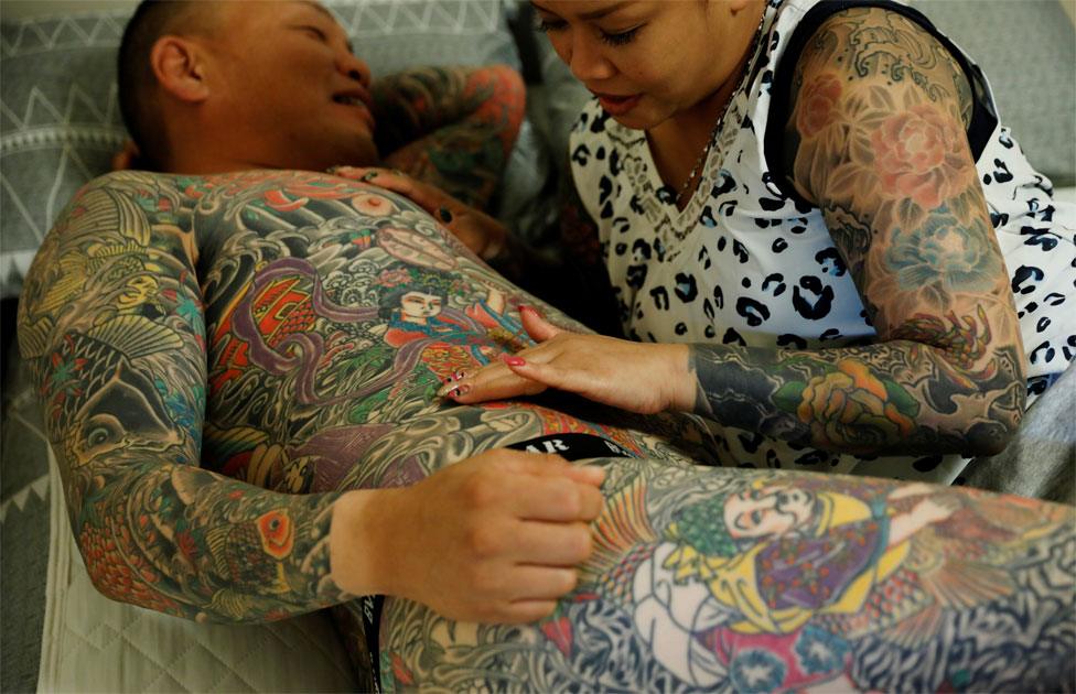 A woman with lots of tattoos touches a tattoo on the torso of her husband