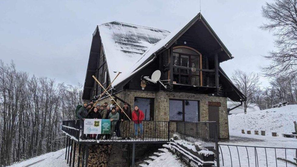 Wooden house on the side of a snow-covered mountain. A group of people is standing behind a Metta sign