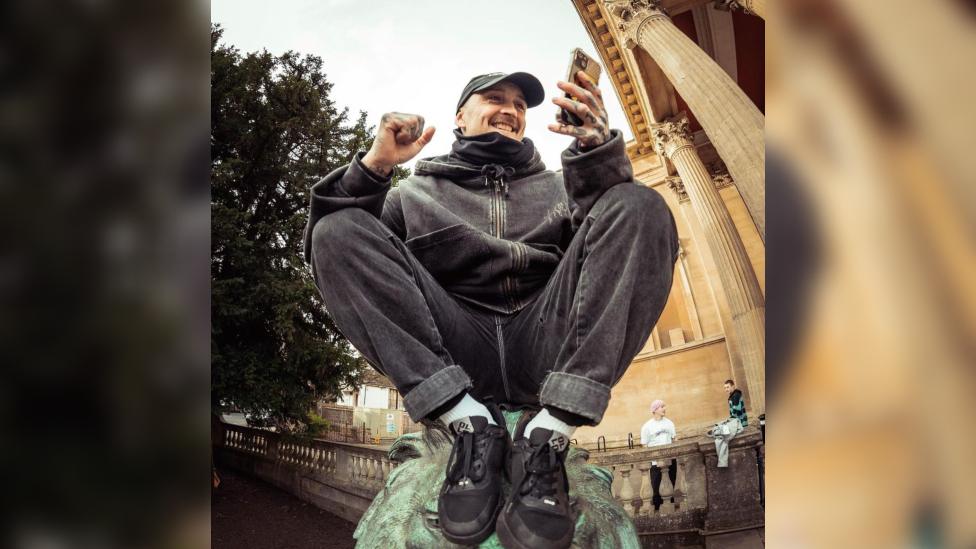 Danny Pierce sitting on top of a fountain statue and smiling at his phone