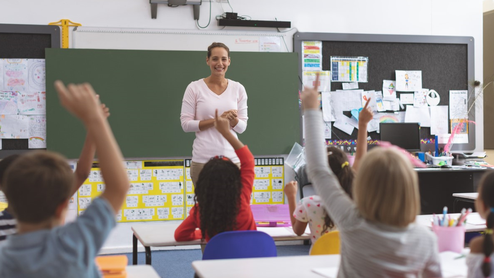 Rear view of school kids raising hand while they are sitting on their chair with their teacher in front of them with a green board in background