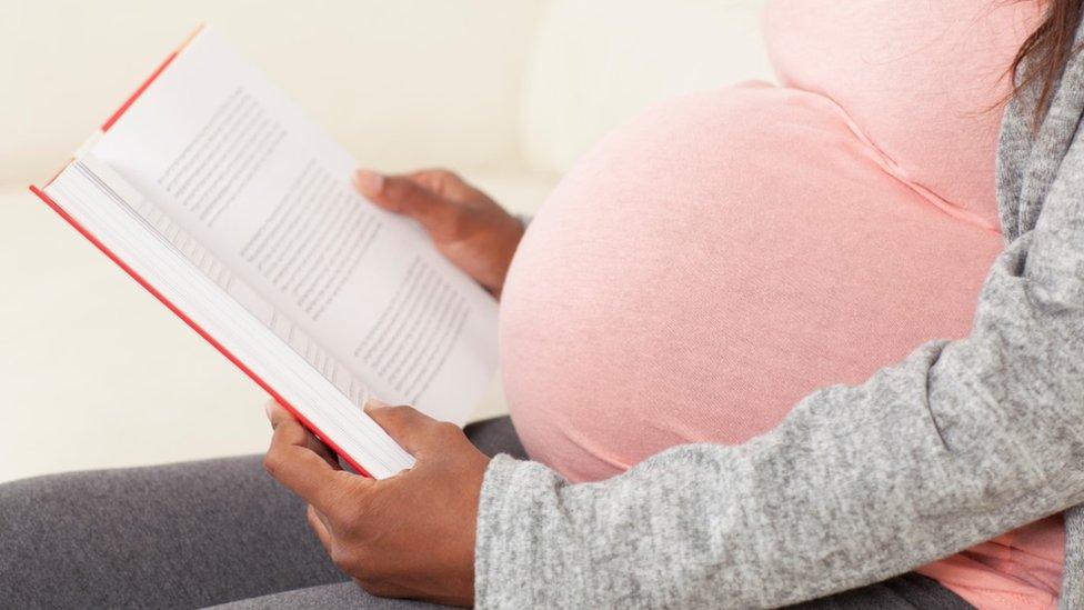 Close up unrecognizable pregnant woman in pink t-shirt reading a book, sitting on sofa.