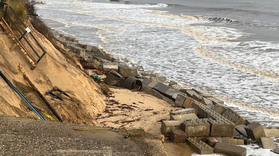 Concrete blocks scattered on beach near crumbling cliff.