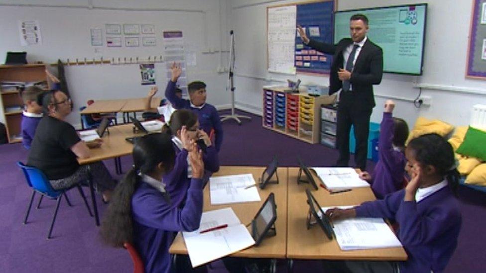 A group of schoolchildren using iPads in a classroom learning from a teacher.