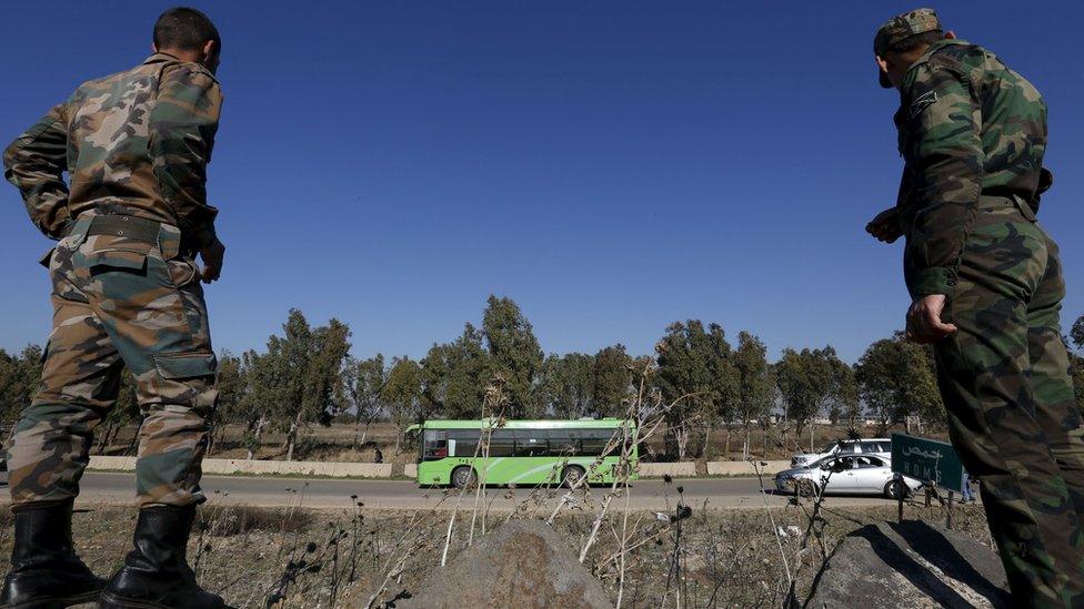 Syrian army soldiers look on as buses carrying rebel fighters leave the al-Wair district of Homs as part of a local truce agreement (9 December 2015)