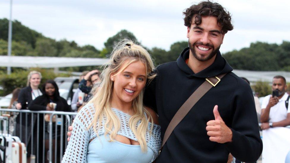 Jess Harding and Sammy Root are seen arriving back in the UK on August 2, 2023 in London. Jess, a blonde white woman, has her long hair in a half-up style. She wears a blue knitted cropped top over a strappy top in the same colour. She has blue eyes and smiles at the camera. Sammy is to her left - he is a white man about a foot taller than Jess. He dark brown curly hair which is cut to just above his ears and has a trimmed dark bears. He wears a black hoody and a brown cross-body bag and smiles at the camera as he gives a thumbs up. the pair are pictured outside at the airport - behind them a small crowd it waiting behind metal barriers.