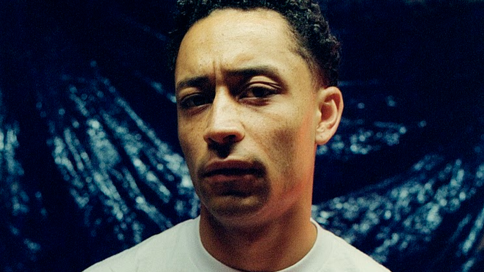 Loyle Carner standing looking at the camera wearing a white T-shirt