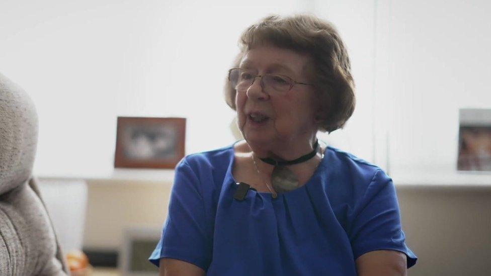 Kathy Jones, a woman with short hair and glasses and wearing a purple top, sitting in her living room in the block of apartments.