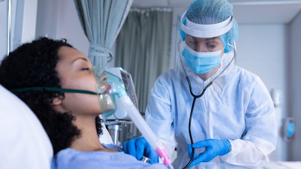 A doctor in PPE with a stethoscope examines a patient receiving oxygen through a ventilator