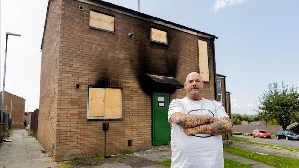 Man standing outside a house which has clear fire damage. The windows have been boarded up.