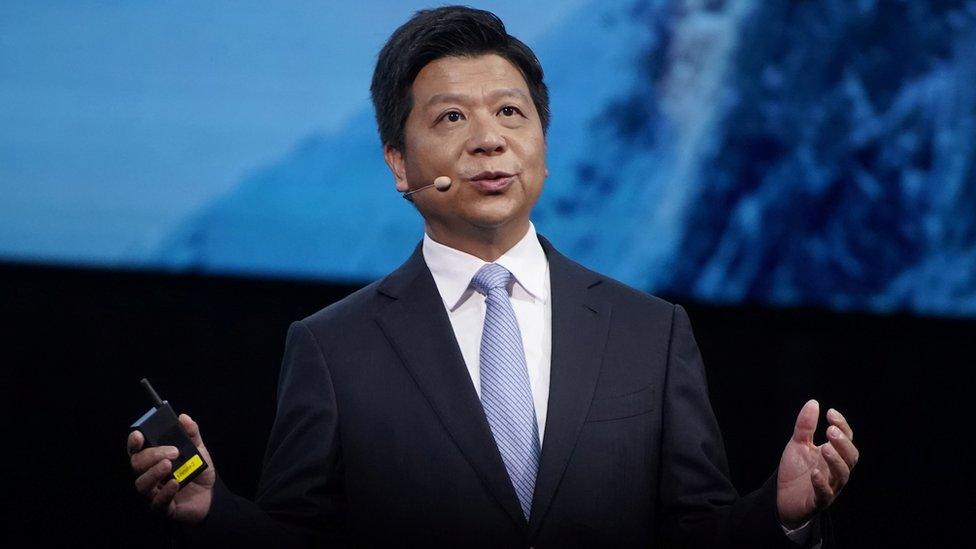 Guo Ping stands in front of a diffused blue photo on a conference stage, arms outstretched while speaking