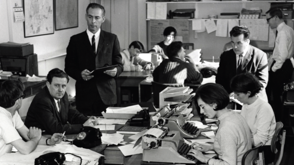 A black and white photo of Cecil Taylor standing in the newsroom among colleagues