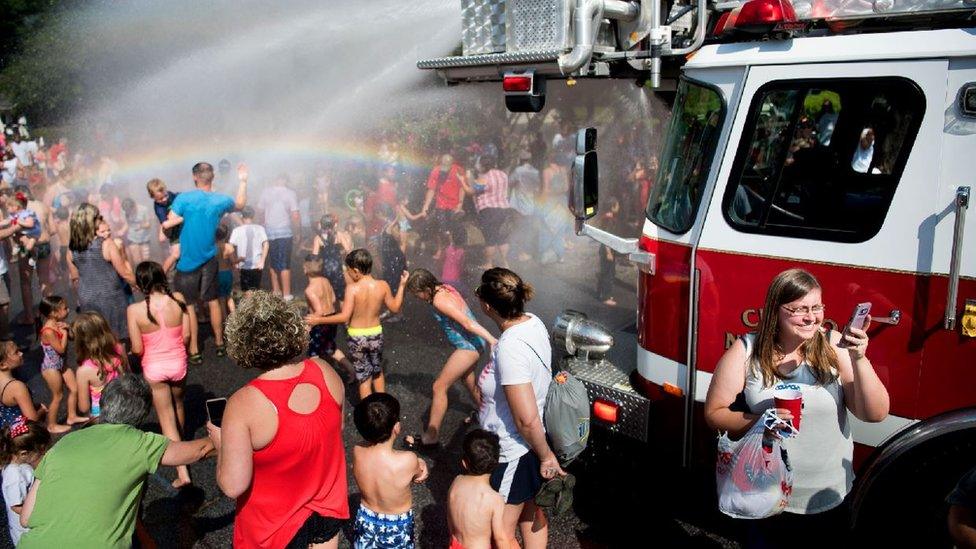 Children and adults pictured playing in spraying water by a truck in the sunshine