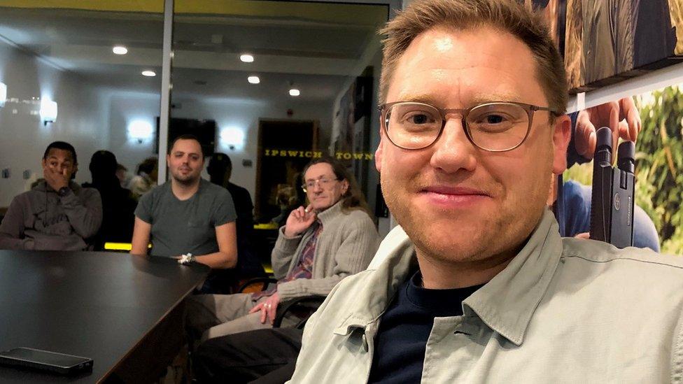 A man with glasses sits in a room with several other men around a table