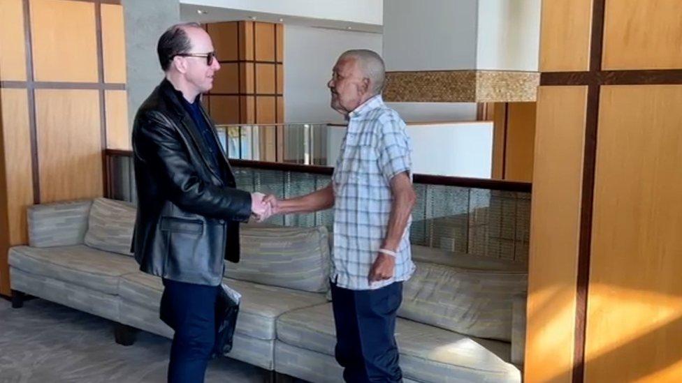 Mark Randolph Dyer (left) is dressed all in black. He is shaking hands with Nizamodeen Hosein