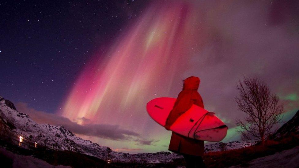 Snow capped mountains with red glow of northern lights and surfer carrying board