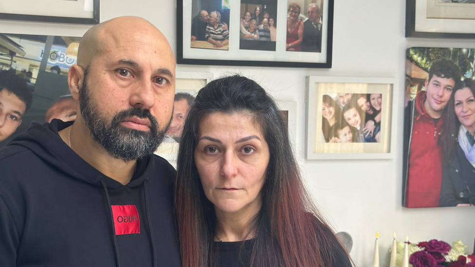 George and Areti Nicolaou in front of a wall of family pictures