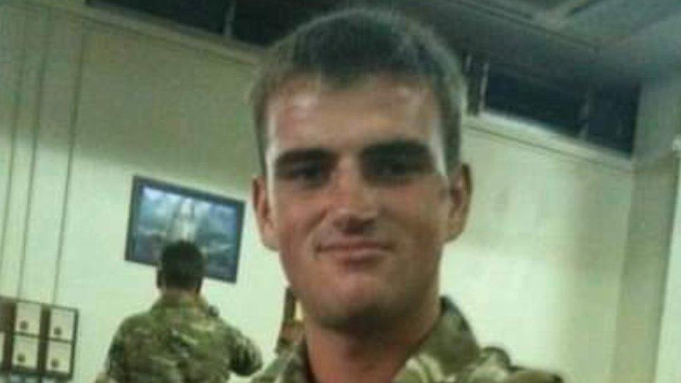 A young man in military camouflage smiles at the camera