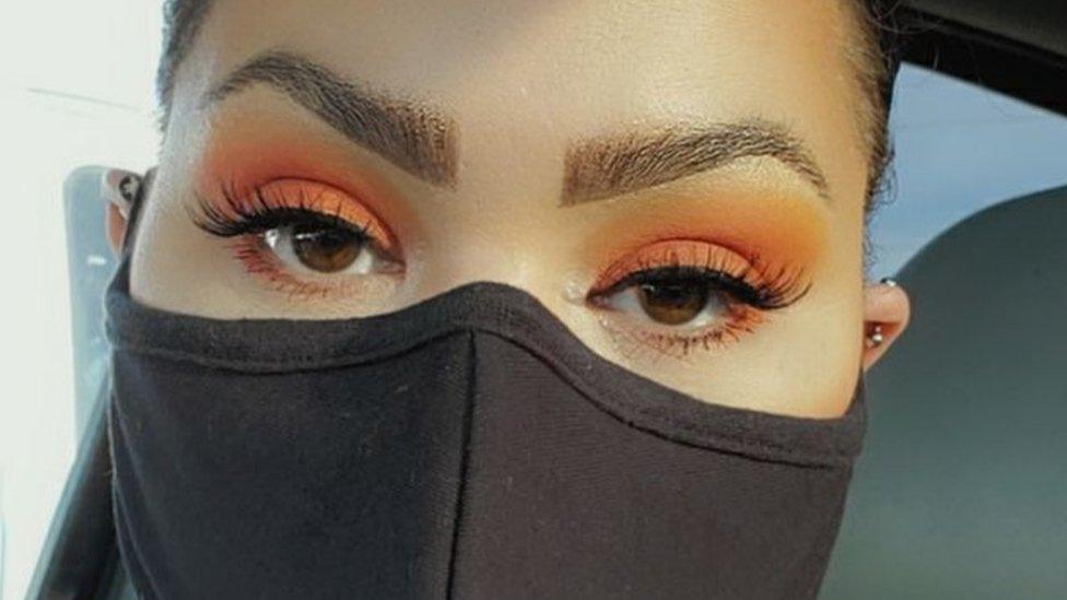 Melina Basnight wears a face mask and a strong eye makeup look, with orange shadow and long false lashes