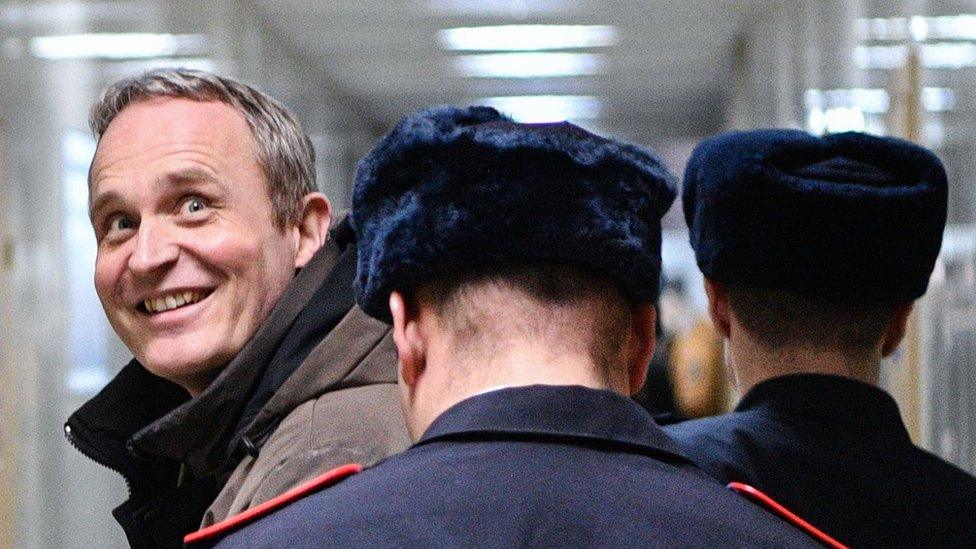 Dennis Christensen is led down a corridor by uniformed police, but looks over his shoulder to smile cheerfully at photographers