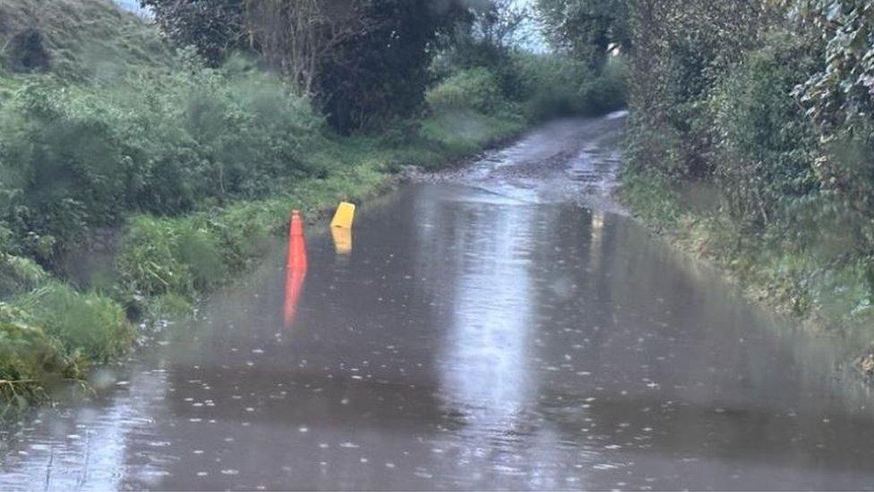 Flooded country lane with two traffic cones peeking out of the top of the muddy water, raining.