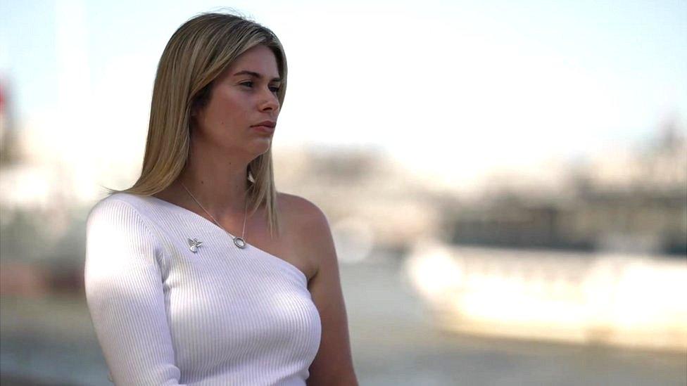 An image of Lauren, a young woman with light hair wearing a white top and an angel brooch.